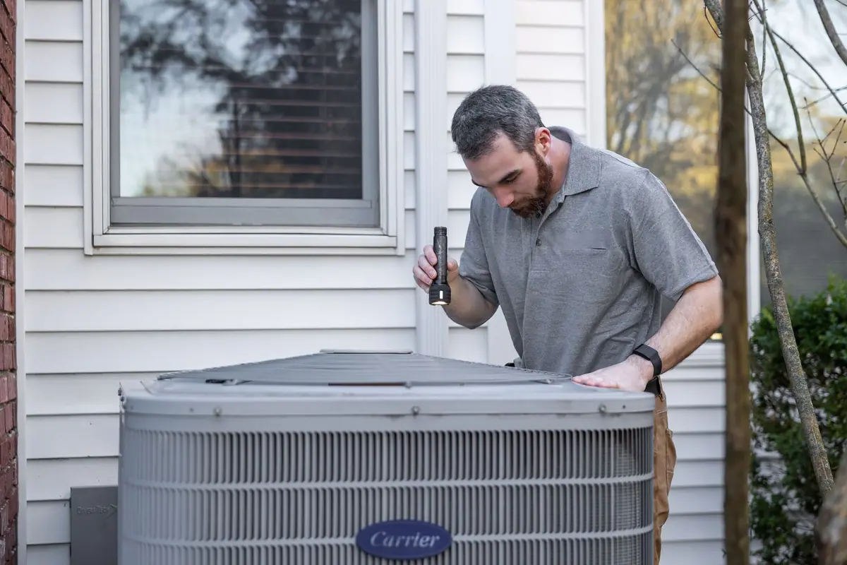 A technician checking the wiring inside the panel of an outdoor condensing unit beside a house.