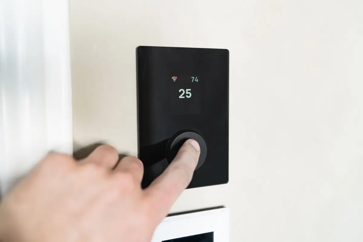 A hand turning the dial on a slim black smart thermostat mounted on a pale wall.