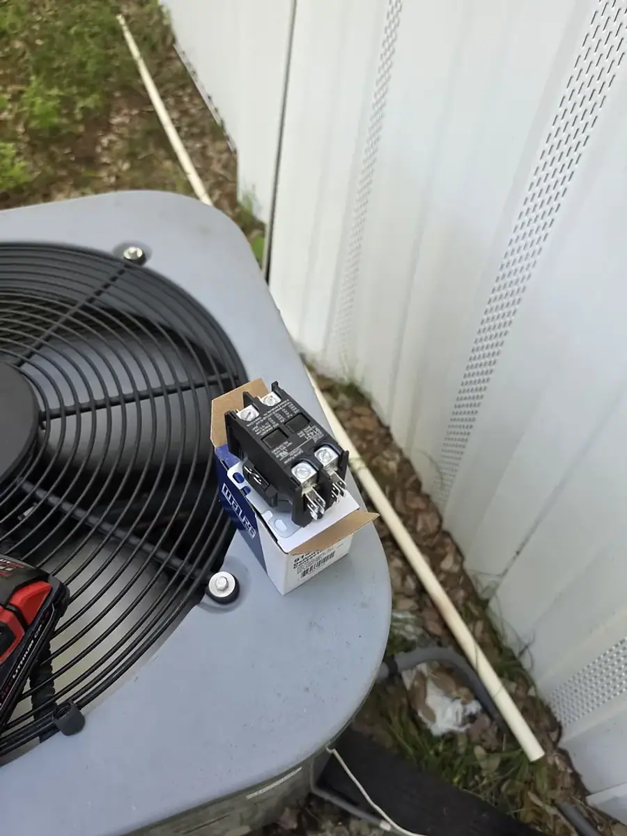 A replacement contactor in its box resting on a condenser beside a house wall.